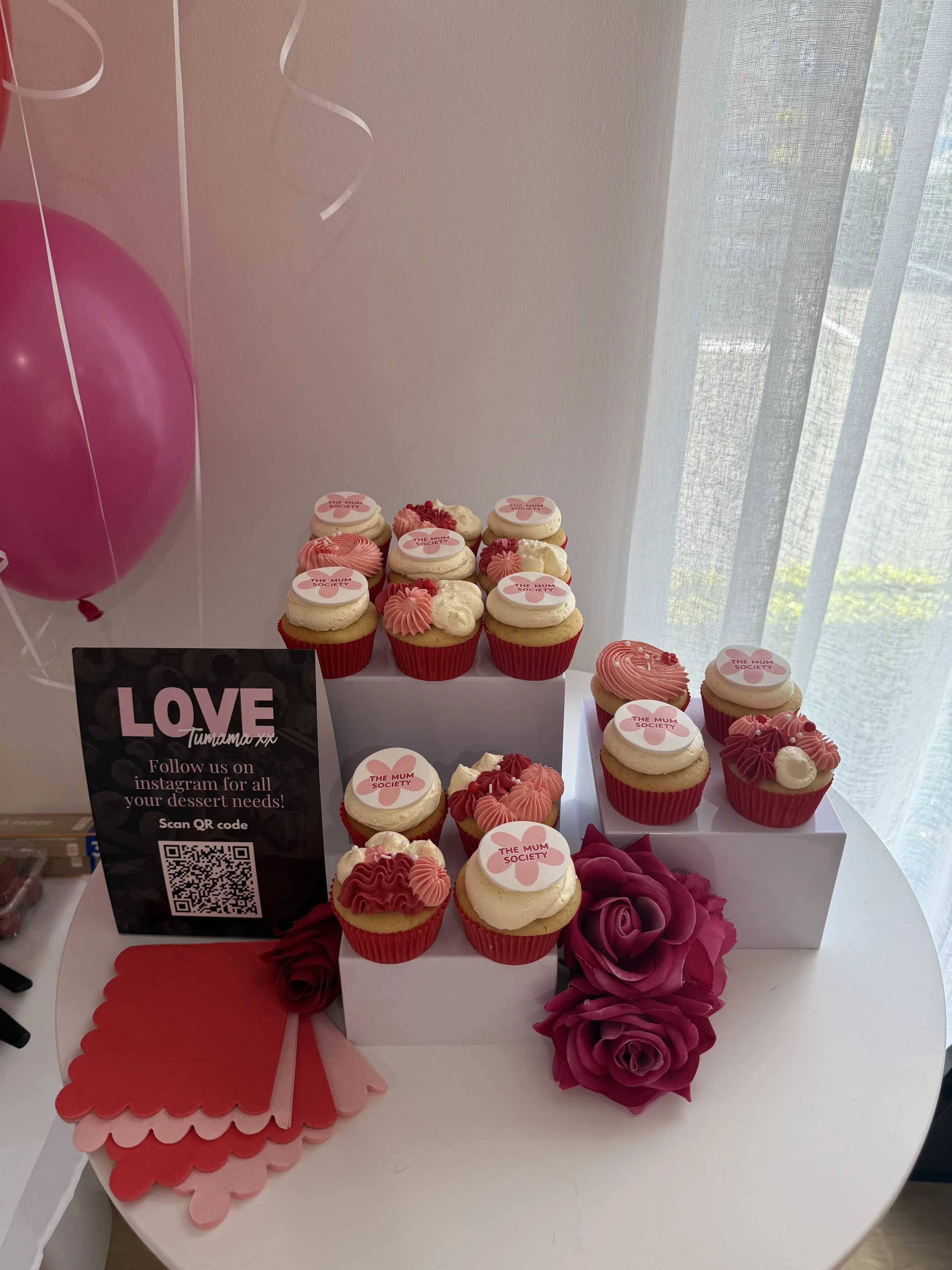 Cupcakes decorated with pink, white, and red frosting, some with edible toppers reading "The Mum Society," on a display table with pink balloons, purple paper roses, and pink and red paper napkins.