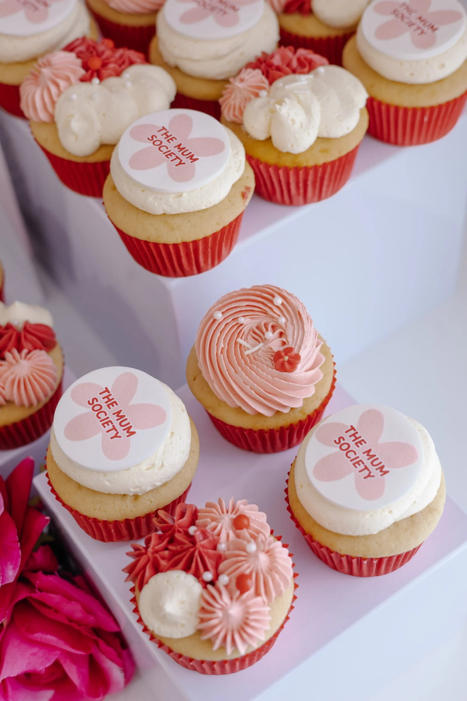 Cupcakes decorated with pink and white frosting, some with edible toppers that say 'The Mum Society,' and others with floral decorations, arranged on white display stands.