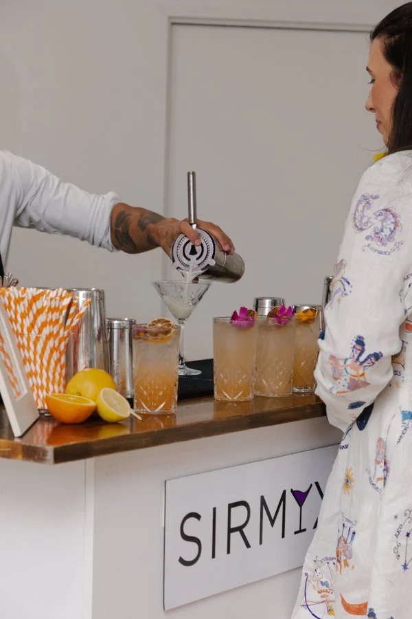 A bartender is pouring a cocktail from a cocktail shaker into a glass with a lemon slice garnish. Several other cocktails with flower garnishes are on the bar, and a woman wearing a white robe with colorful prints is watching.
