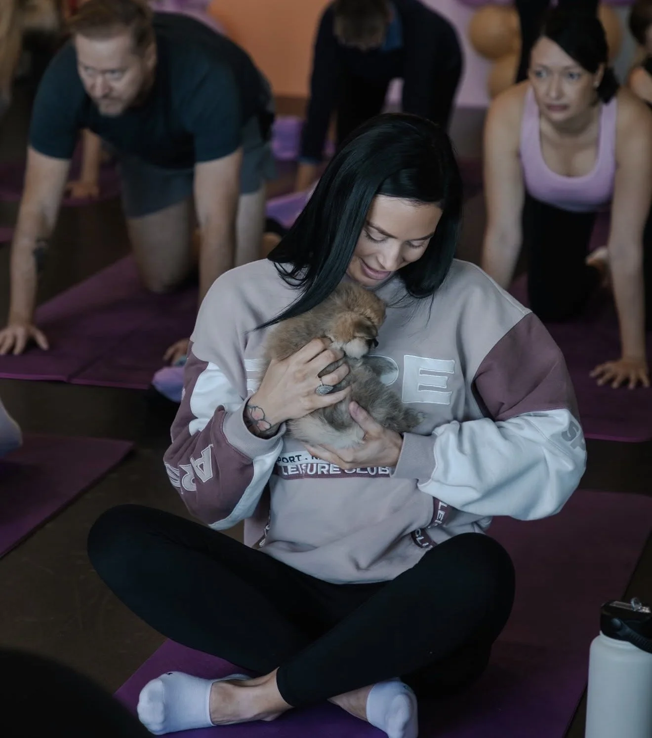 A woman sitting cross-legged on a yoga mat in a group exercise class, holding a puppy close to her chest while surrounded by other participants in yoga poses.