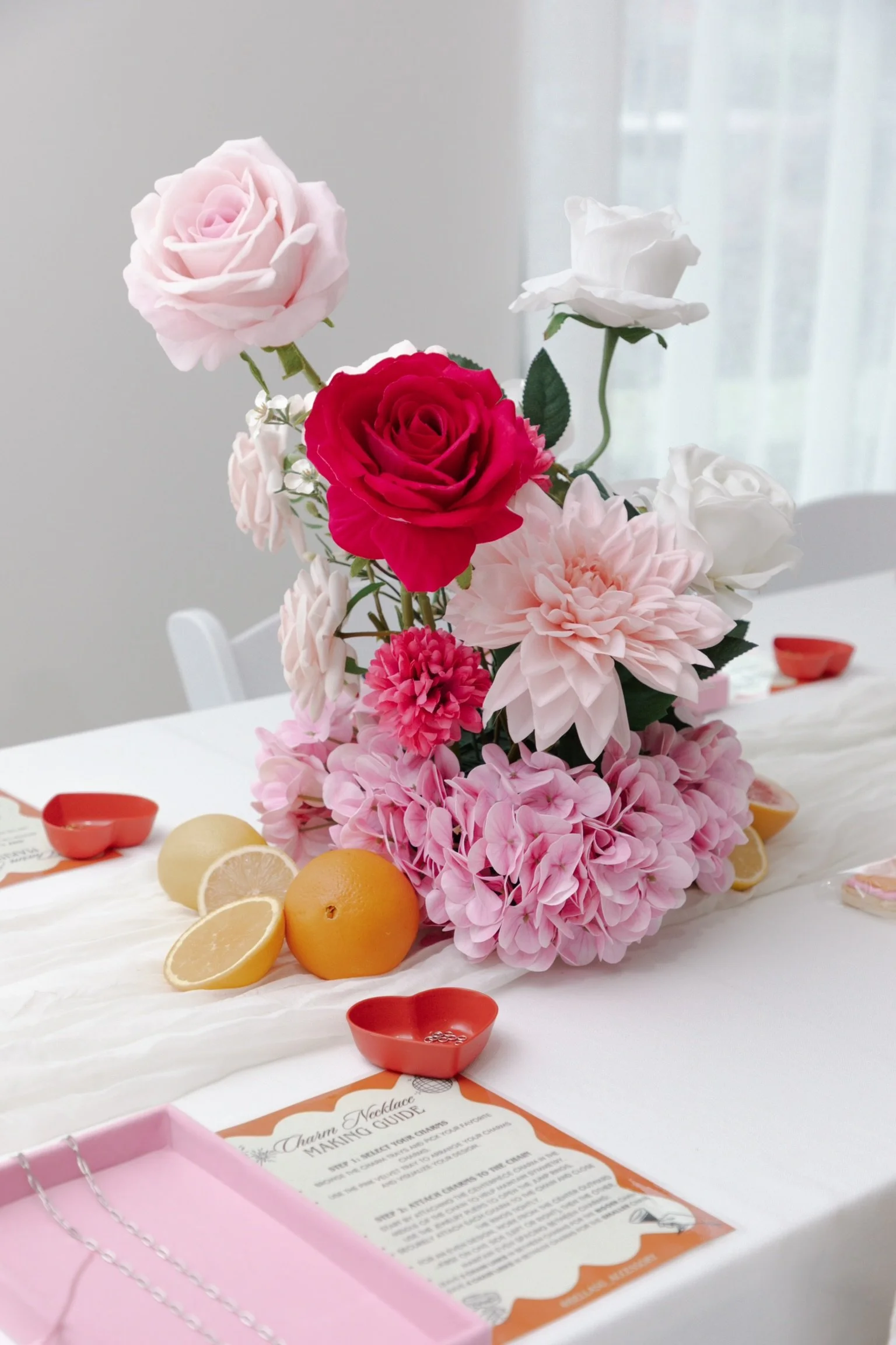 A floral centerpiece with pink, white, and red roses and pink hydrangeas on a white tablecloth, decorated with lemon and orange slices, small red heart-shaped dishes, and event programs.