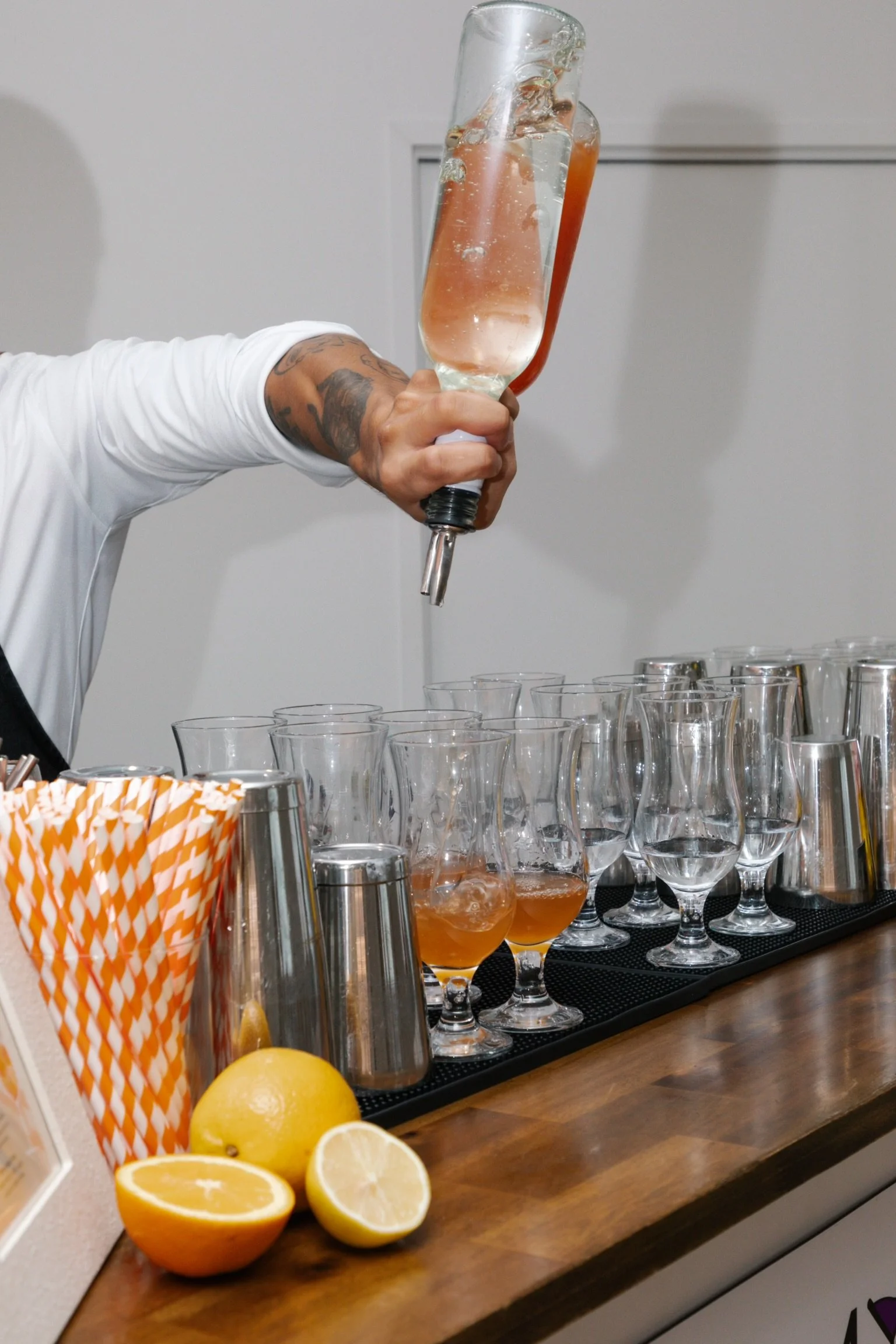 Person pouring a pink cocktail from a bottle into glasses on a bar counter, with lemons and striped straws in the foreground.