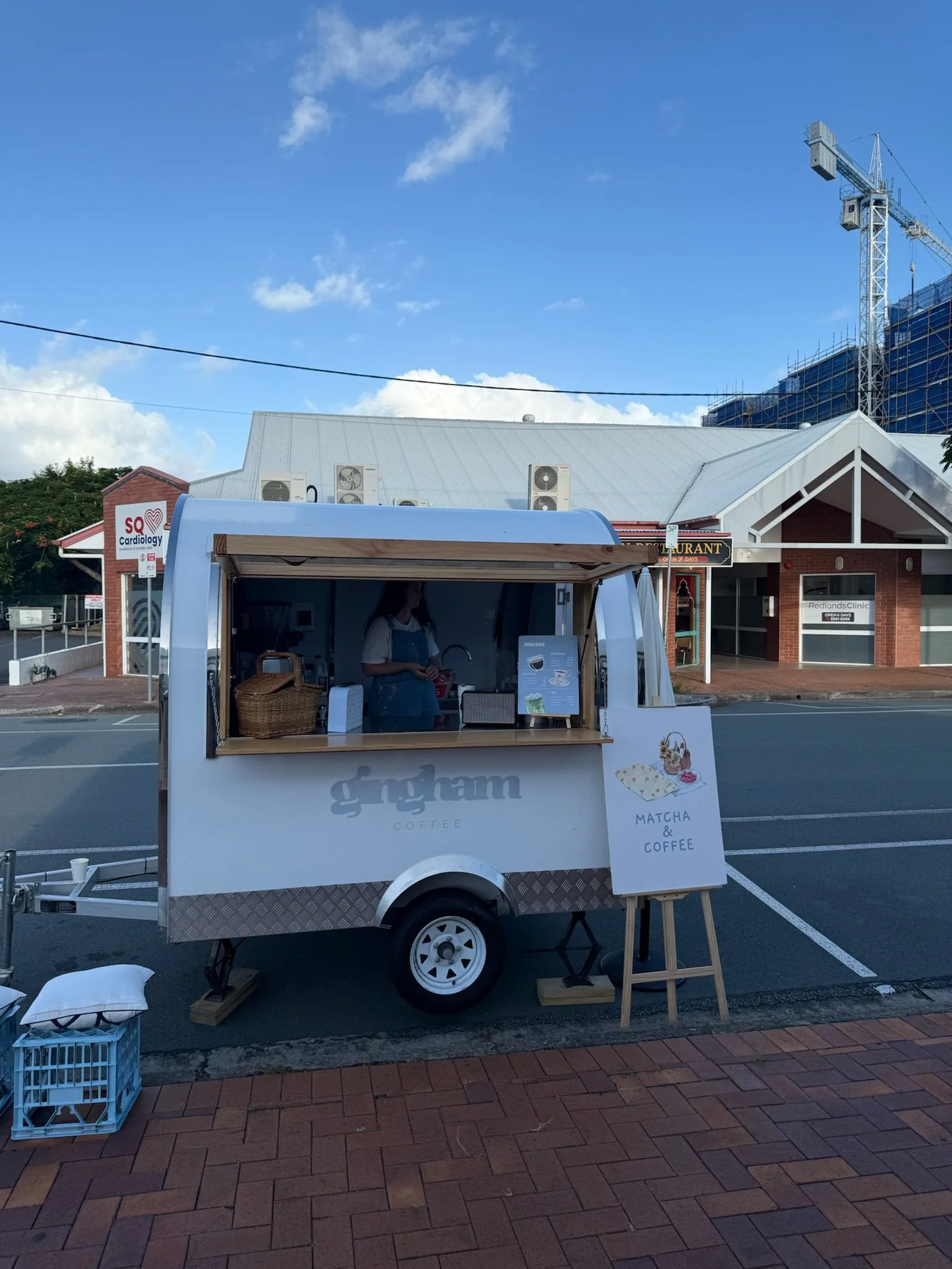A small food truck selling matcha and coffee parked on a street. The truck has a sign on a small easel with an illustrated matcha and coffee drink, and a person inside wearing a blue apron. Buildings and a construction crane are visible in the backgr