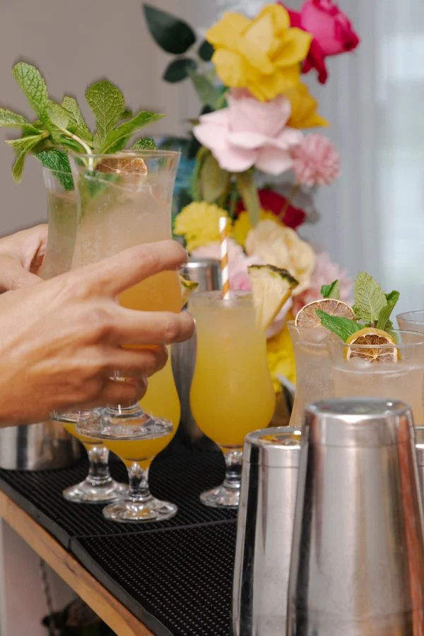 A hand reaching for a tropical cocktail garnished with mint leaves and a lime slice on a bar counter, with other similar drinks and a vase of colorful flowers in the background.