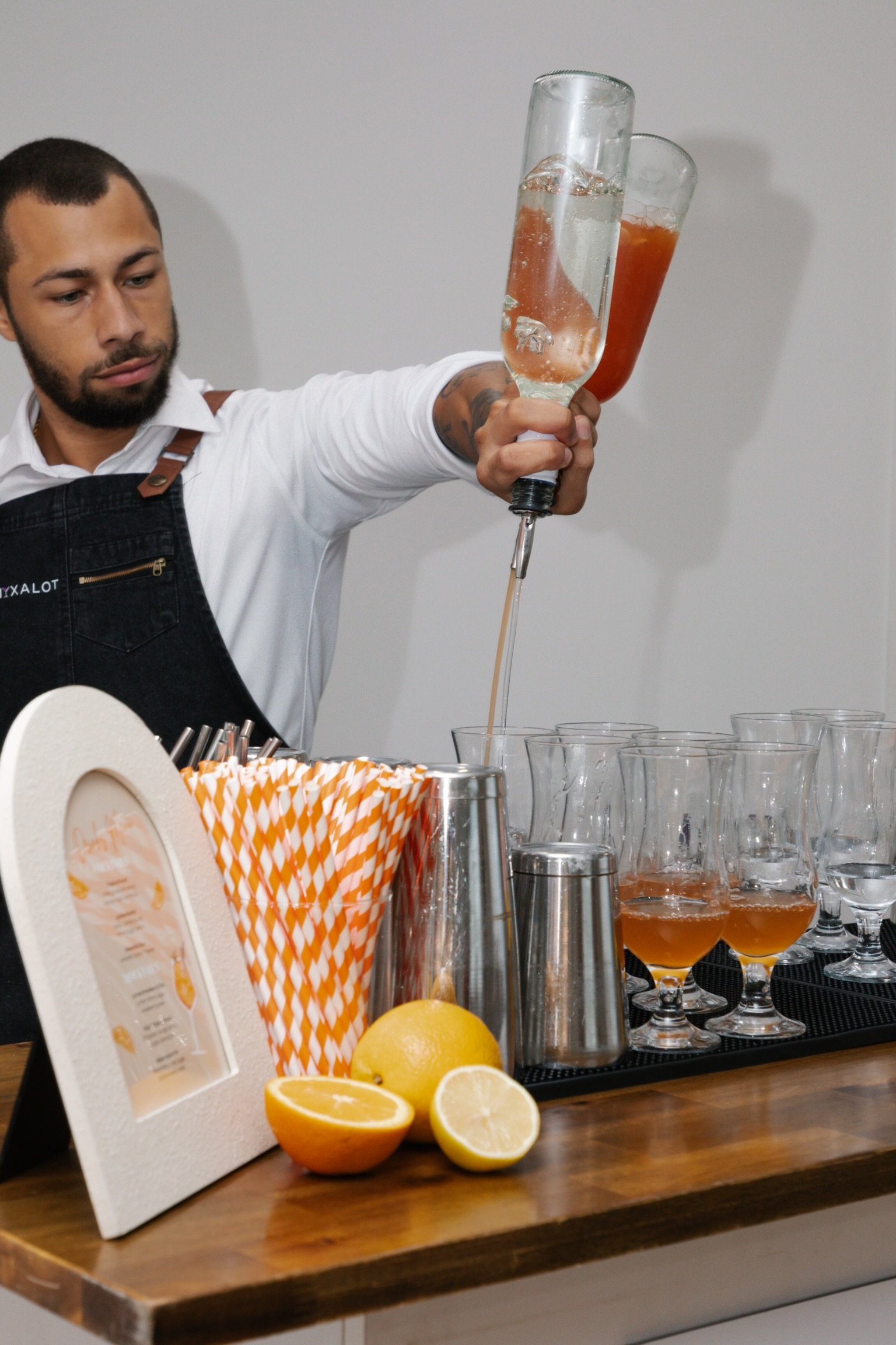 A bartender pouring a cocktail from a bottle into a glass, with several glasses of similar drinks on the bar, citrus fruits, striped straws, and barware on the counter.