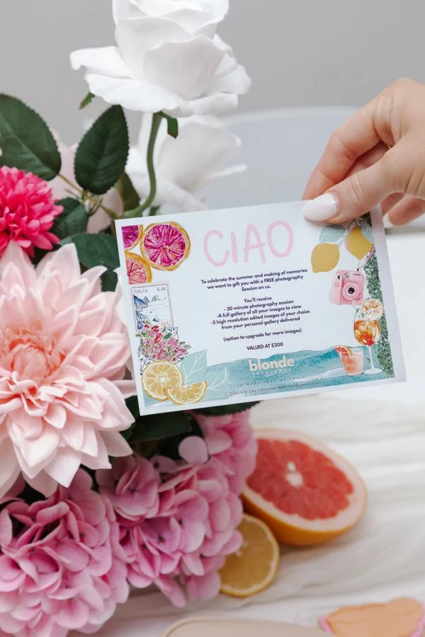 A hand holding a colorful gift certificate or voucher in front of pink and white flowers, with slices of grapefruit and lemon on a light surface.