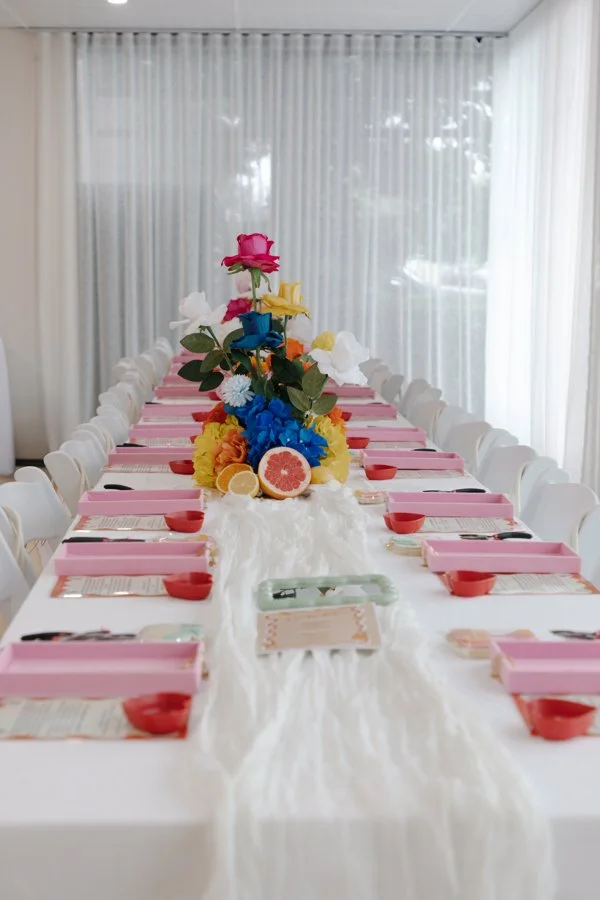 Long banquet table decorated with a white table runner, colorful flower arrangement in the center, pink place settings, and napkins. The background features sheer white curtains.