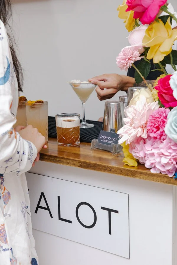 People at a bar with colorful flowers, with drinks including a cocktail and an IPA, and a sign with the word 'ALOT'.
