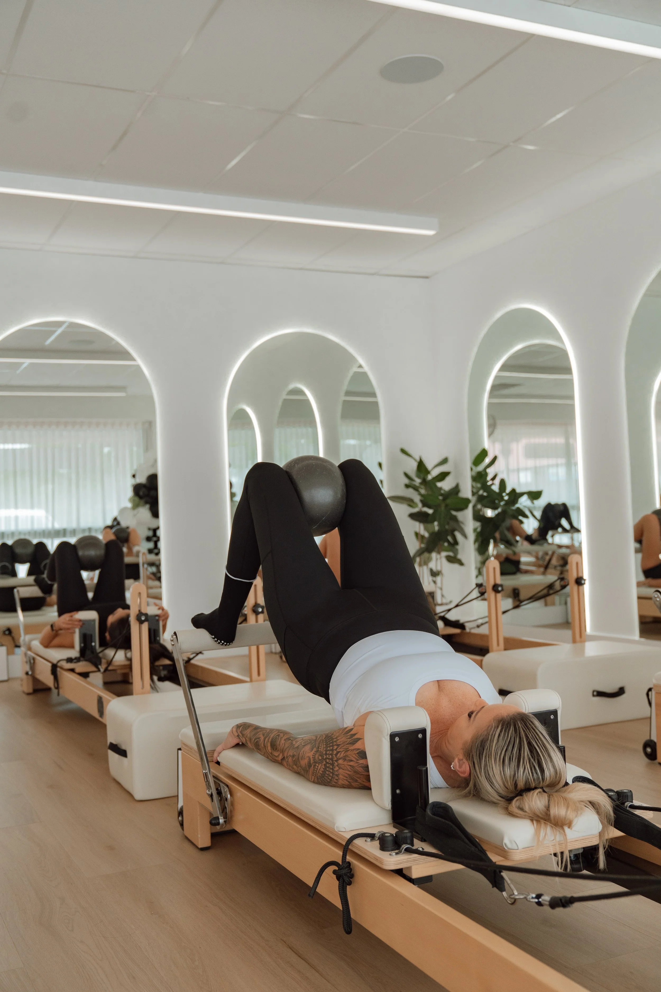 A woman lying on a Pilates reformer machine, performing an exercise with a ball positioned between her legs. The room has white walls with arched mirrors, and other people are exercising in the background.