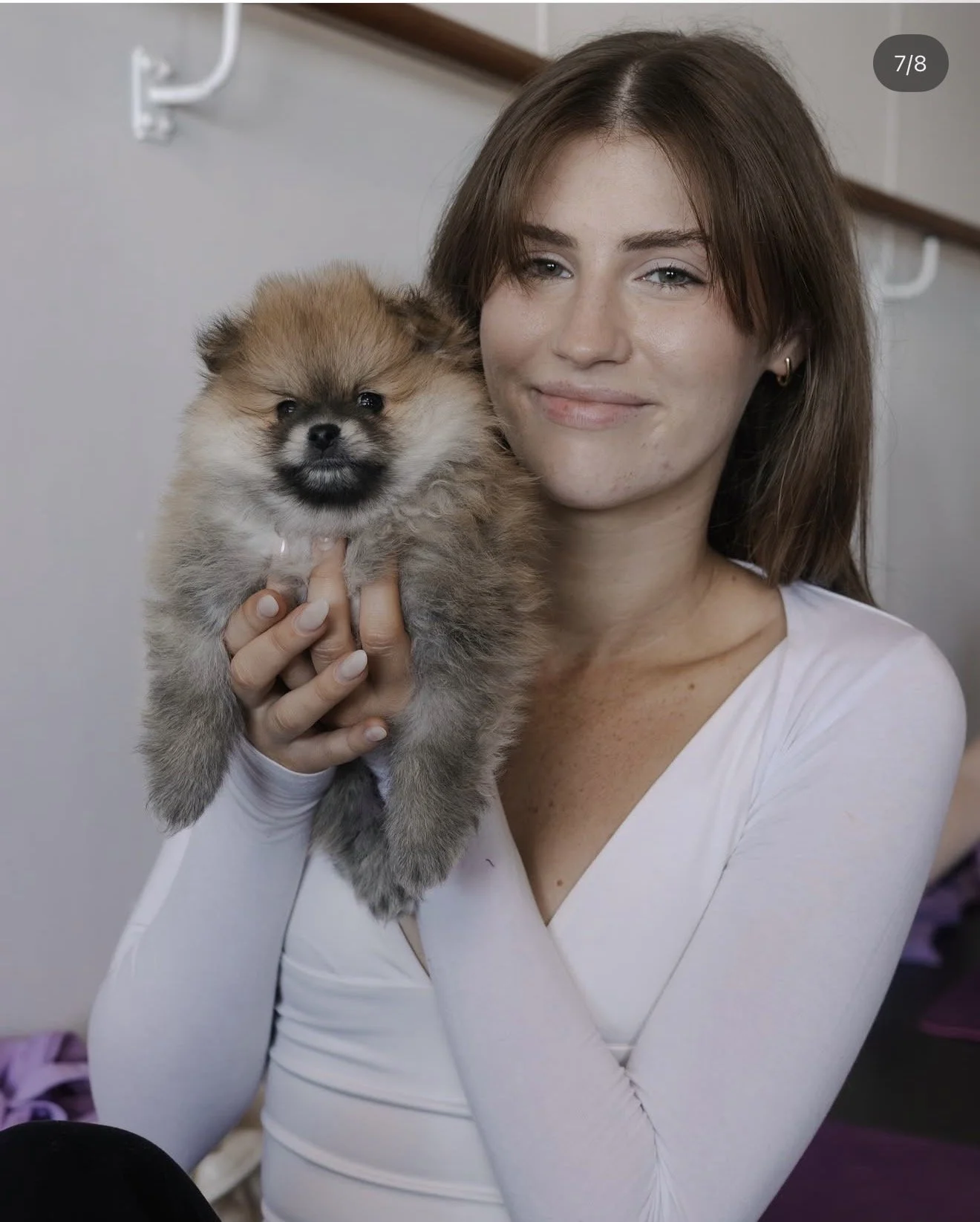 A young woman with light skin and brown hair smiling while holding a fluffy Pomeranian puppy.