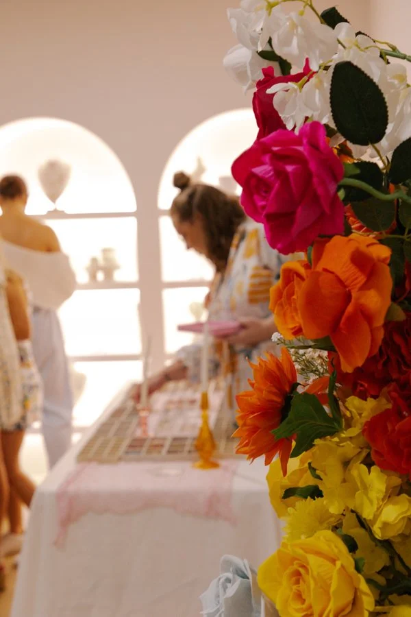 A colorful flower arrangement with white, pink, orange, and yellow flowers in foreground, with blurred people shopping at a display table in the background.
