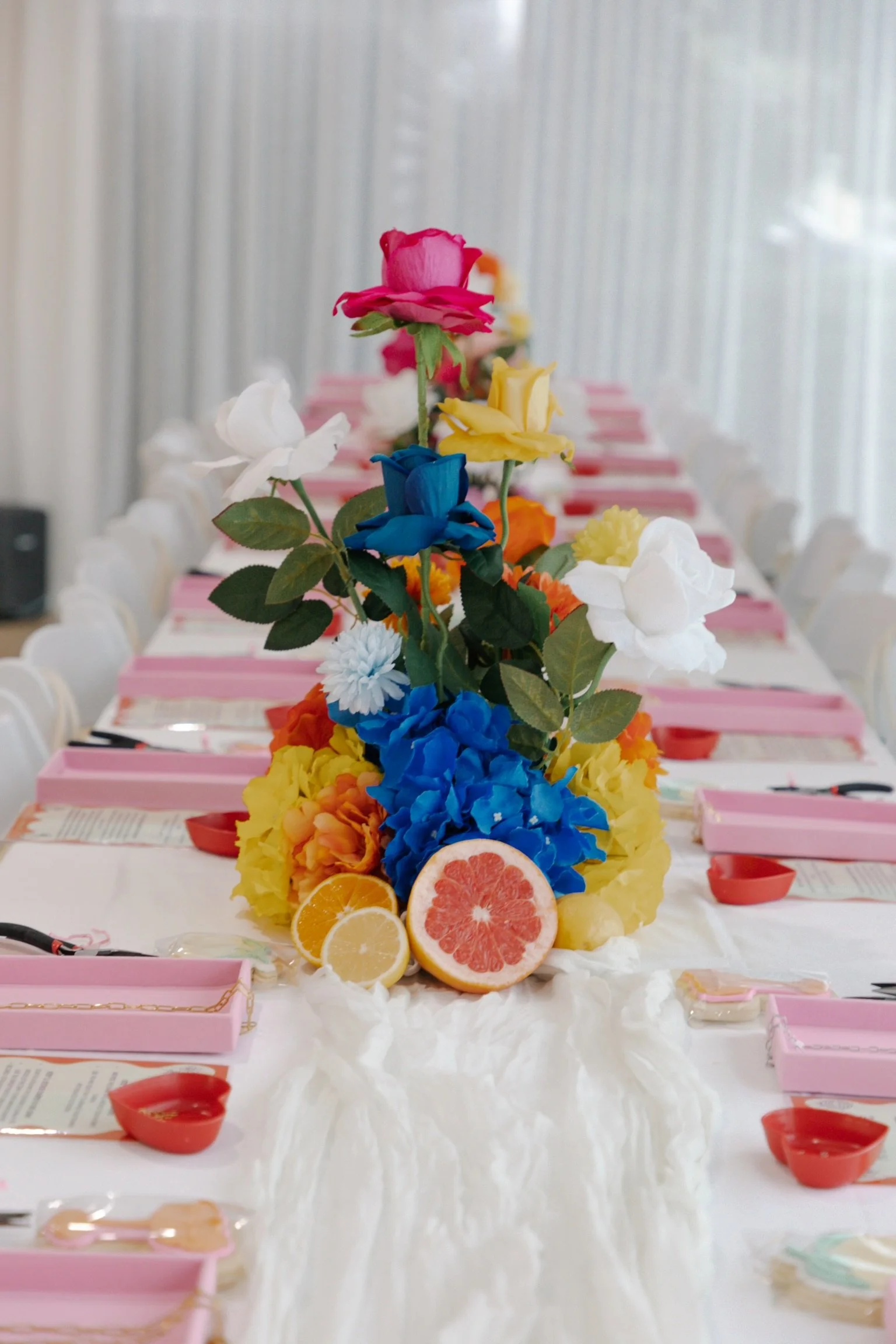 A decorated table with a colorful flower centerpiece, slices of citrus fruits, and pink boxes on a white tablecloth, set for a celebration or event.