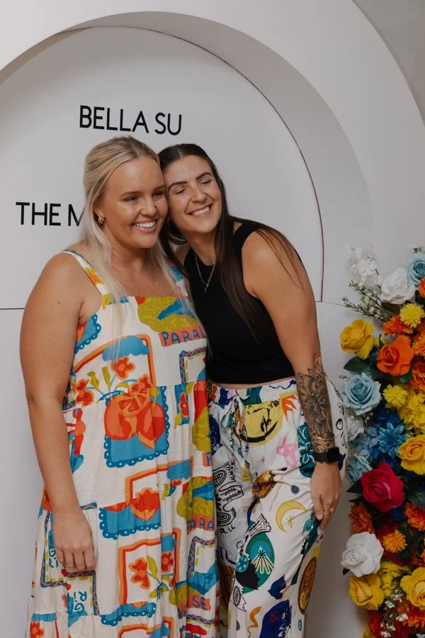 Two women smiling and hugging in front of a white wall with black text and a colorful flower arrangement on the side.