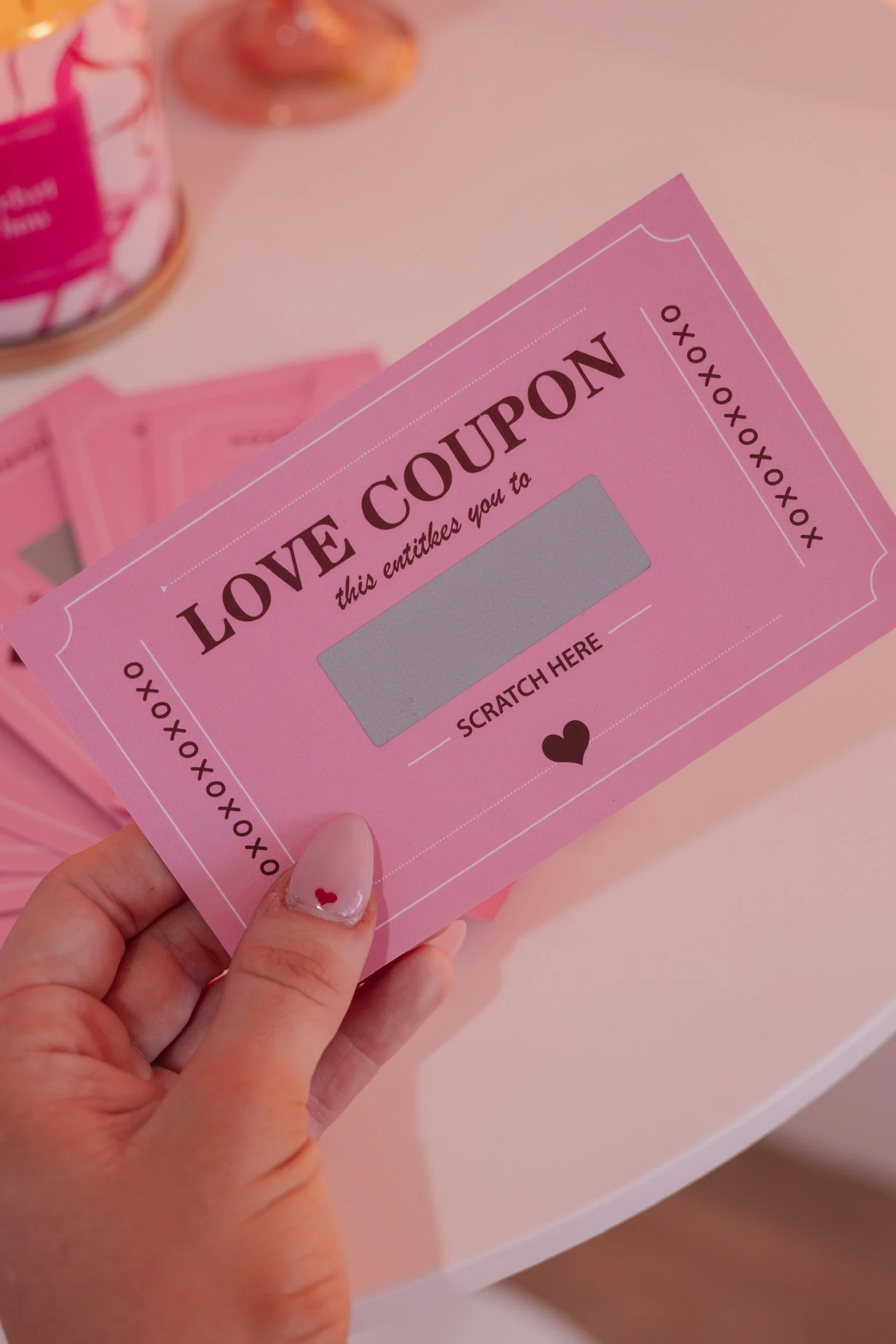 A hand holding a pink love coupon with a scratch-off area, underneath a surface with additional pink coupons and a jar in the background.