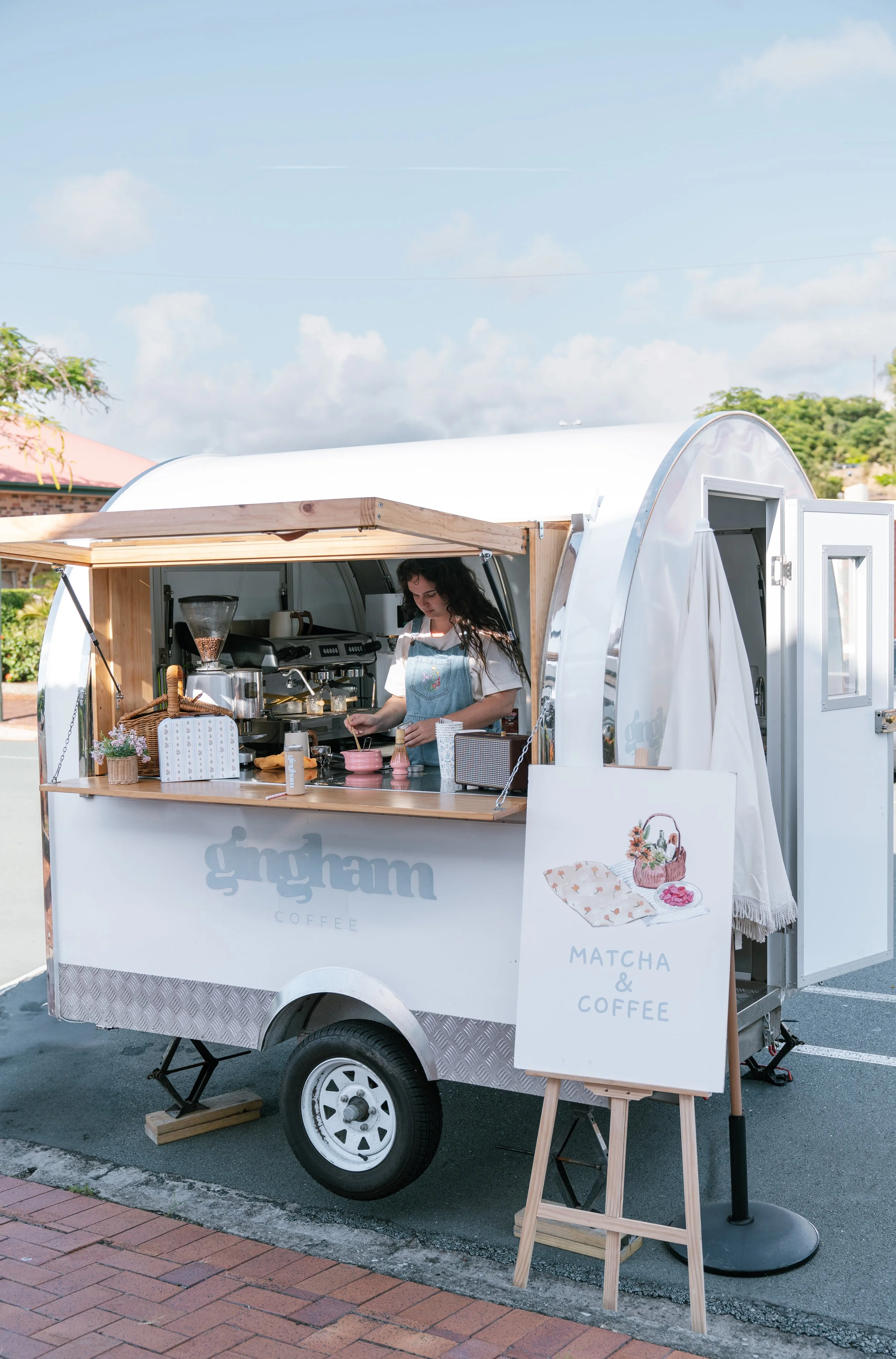 A woman preparing coffee inside a small, white coffee trailer labeled 'grgham'. The trailer has a wooden service window, and a sign outside offers matcha and coffee. The setting appears to be outdoors on a sunny day.