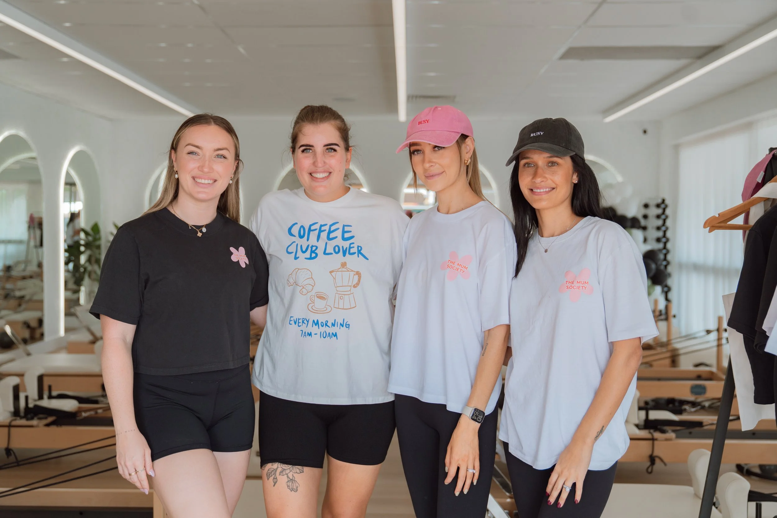 Four women standing together in a fitness studio, smiling at the camera. They are wearing casual workout clothes, with two of them in white T-shirts and two in darker tops. The space has mirrors, exercise equipment, and wooden floor mats.
