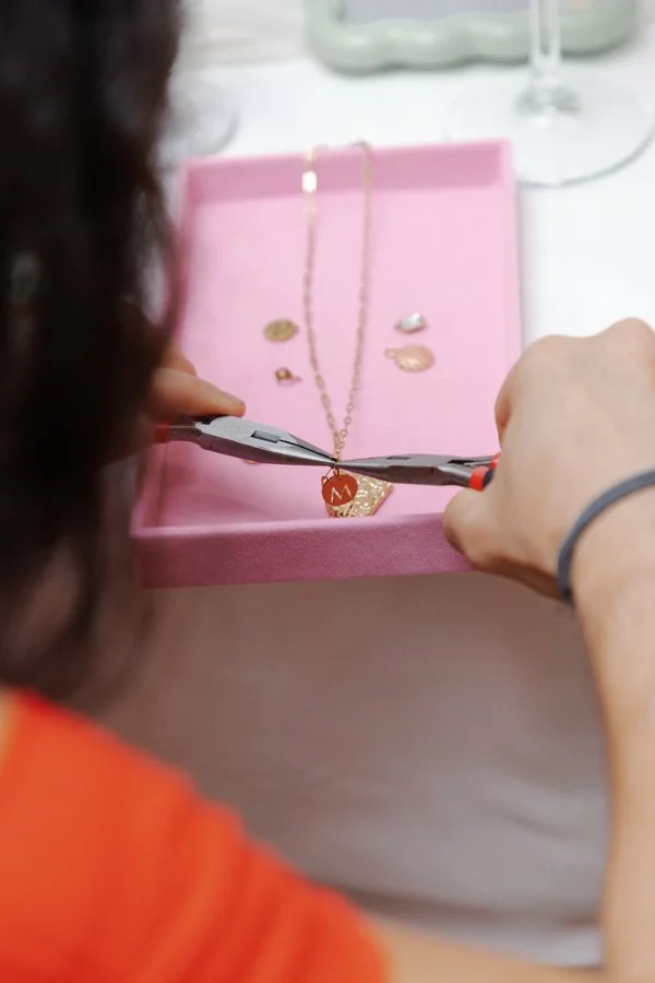 A person is using pliers to adjust or repair a gold necklace with a red tag, placed inside a pink jewelry box with additional jewelry pieces.