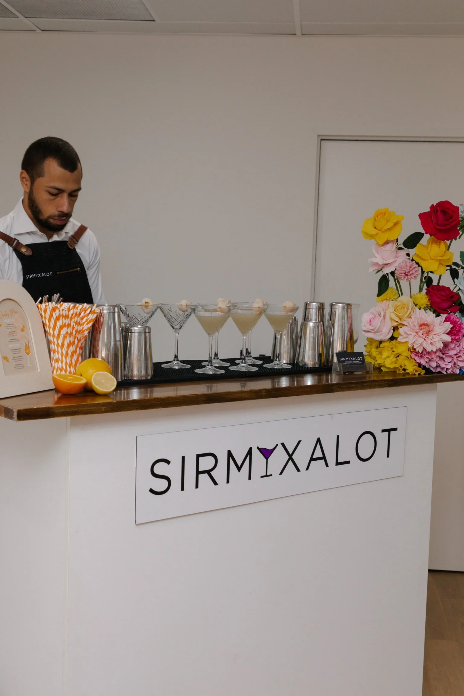 A bartender standing behind a bar with a SIRMXALOT sign, colorful flowers to the right, and martini glasses with drinks on the bar.