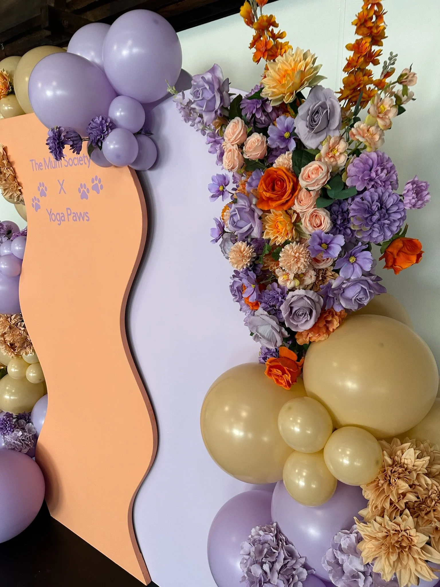 Decorative floral arrangement with purple, orange, and peach flowers, and pastel balloons in purple, beige, and yellow, with a sign that reads 'The Mums Society x Yoga Paws'.