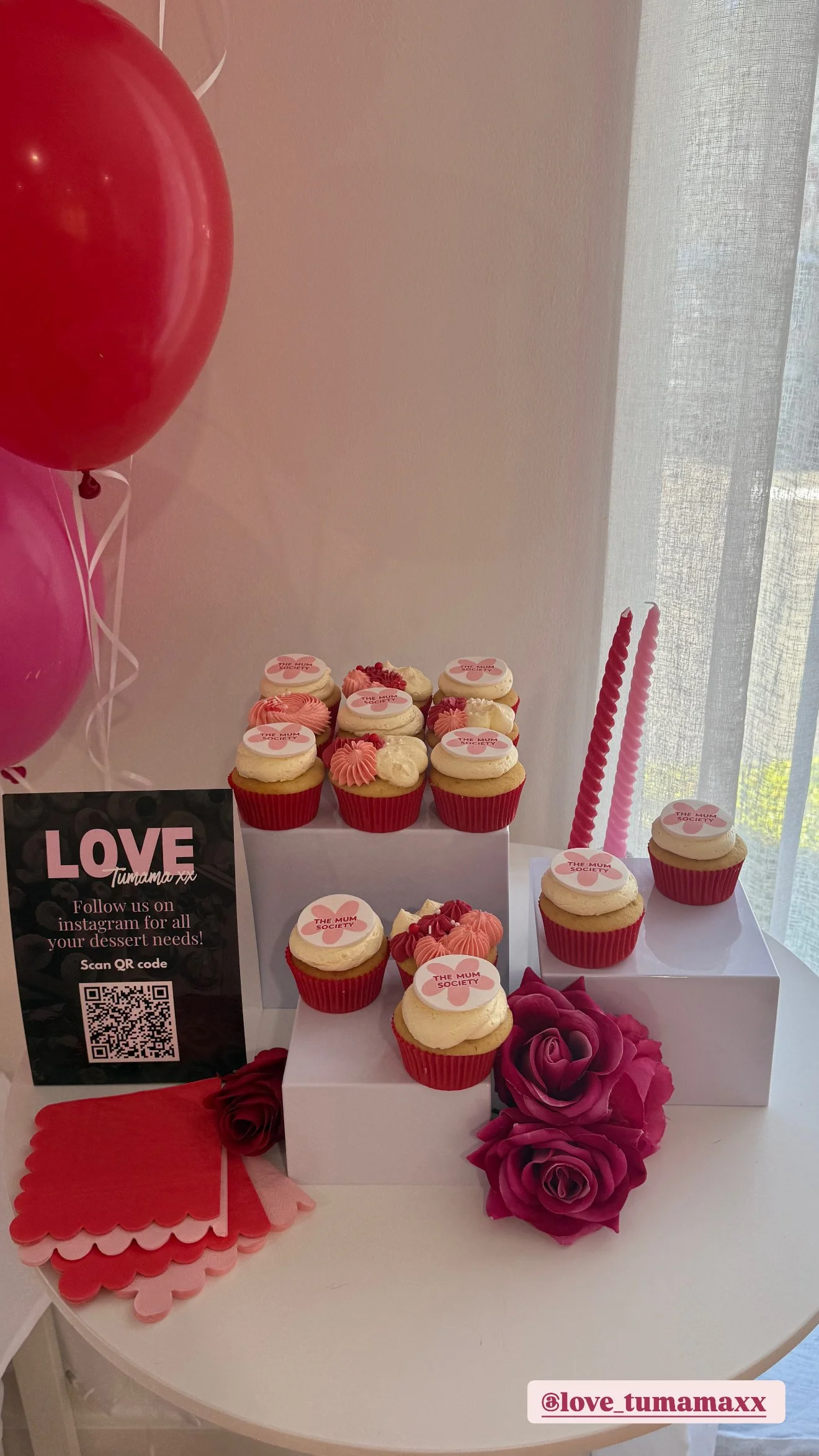 Valentine-themed cupcake display with pink and white frosting, red cupcake liners, decorative pink roses, red balloons, and a black sign with pink and white text promoting an Instagram page for dessert needs.