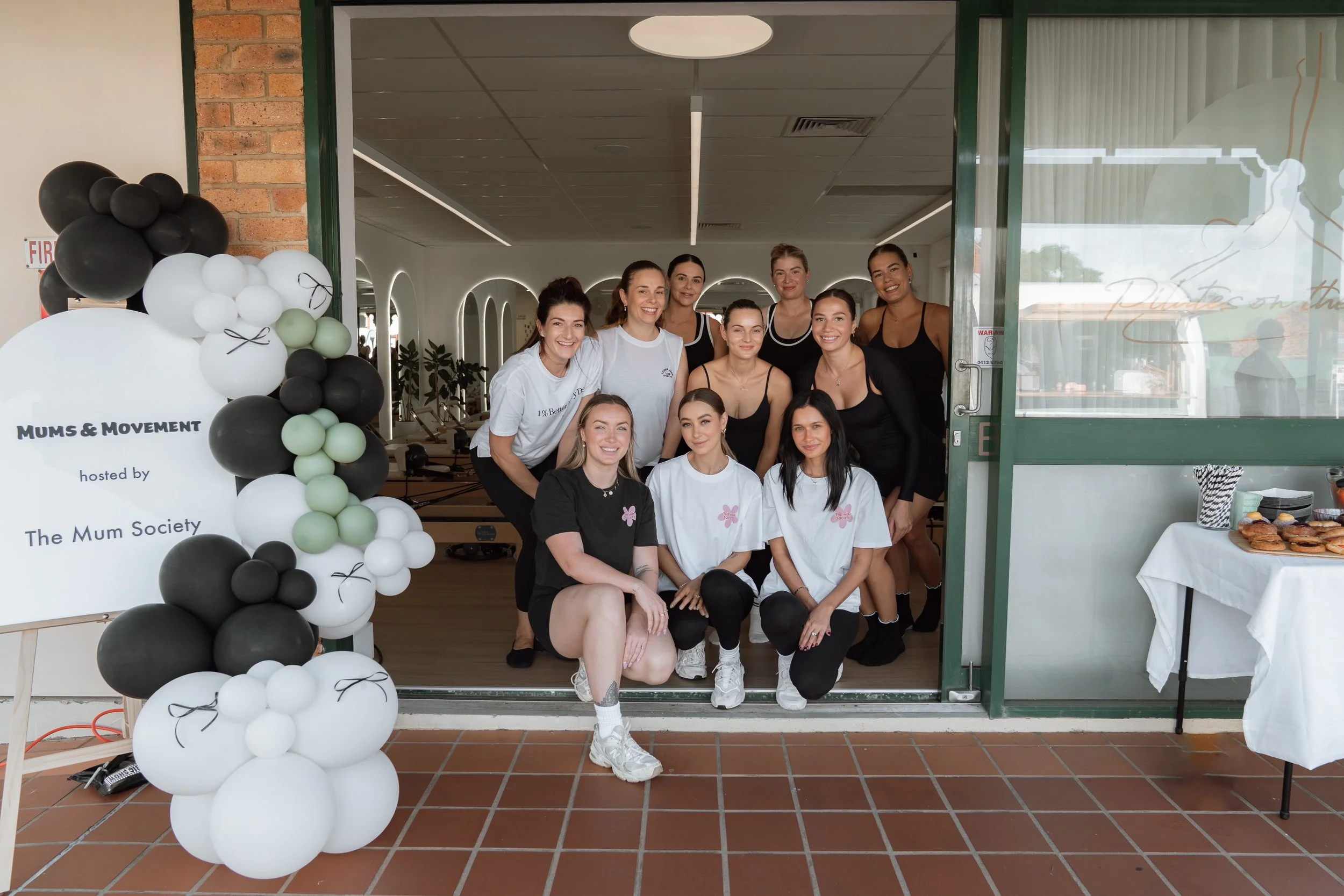 Group of women and girls posing at the entrance of a fitness or dance studio, with a balloon arrangement and a sign that reads 'Mums & Movement hosted by The Mum Society'.