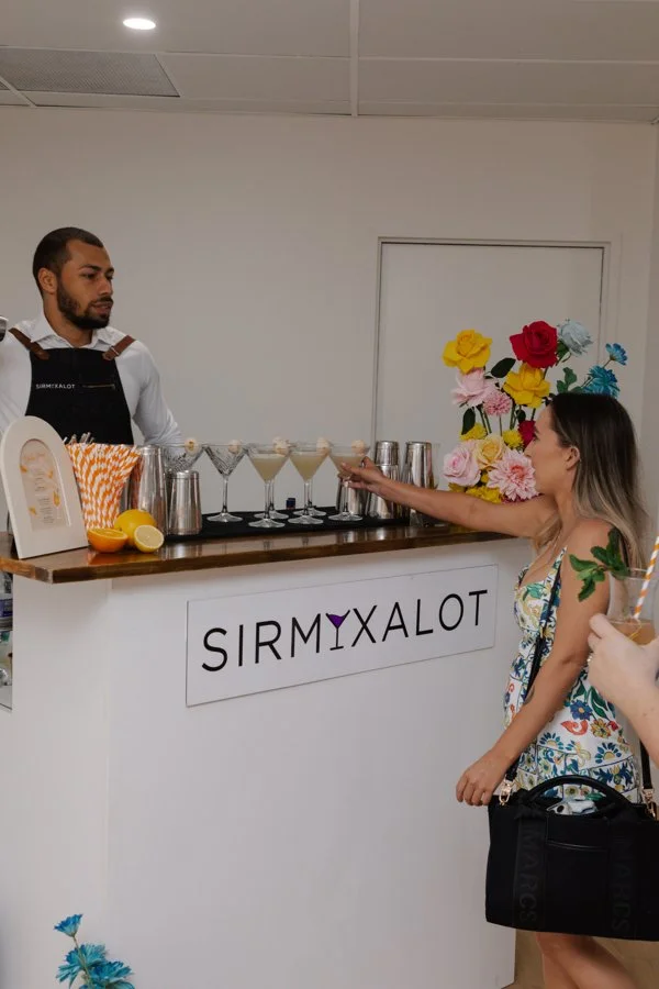 A woman at a bar labeled 'SIRMXALOT' is reaching out to a bartender for a drink. The bar has a wooden top, colorful paper flowers in the background, and several cocktail glasses filled with drinks.