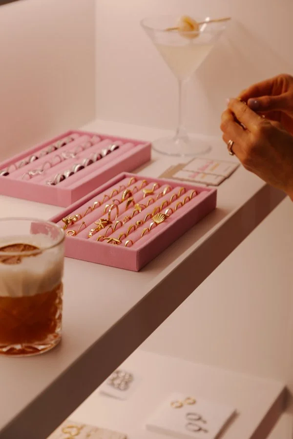 Pink jewelry display boxes with various gold rings, a person's hands holding a ring, and cocktails on a white table.