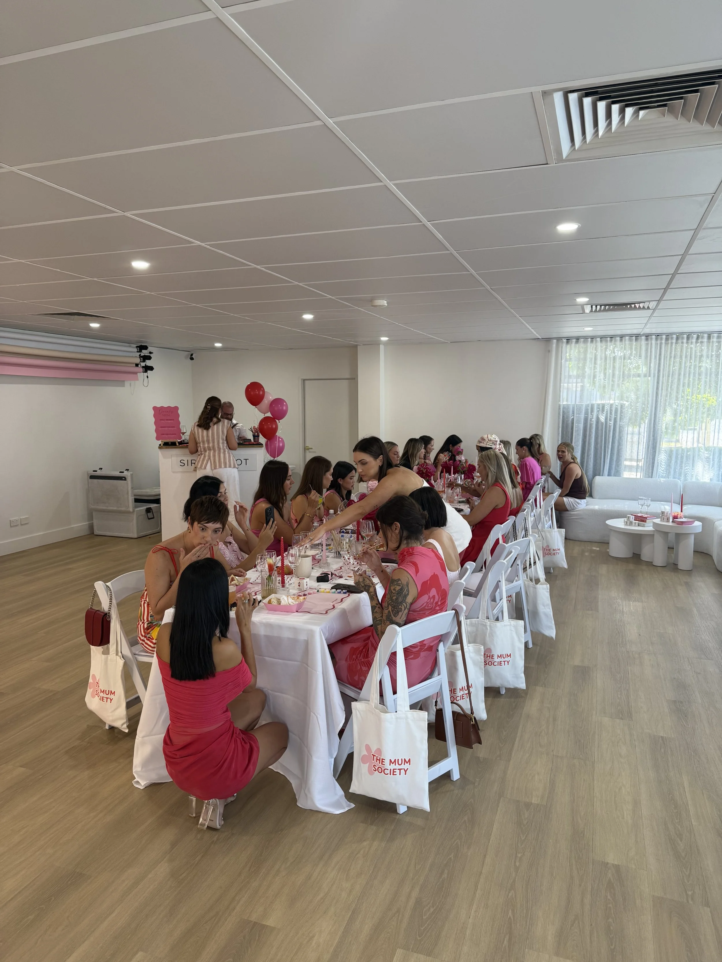 A group of women gathered around a long table decorated with pink and red balloons, pink tableware, and flowers for a celebration at The Mum Society, inside a bright, airy room with white walls, hardwood flooring, and large windows.