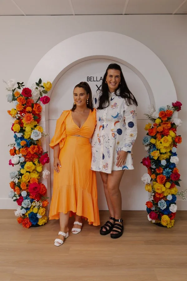 Two women standing in front of a white decorative arch surrounded by colorful flower arrangements, smiling for the photo.