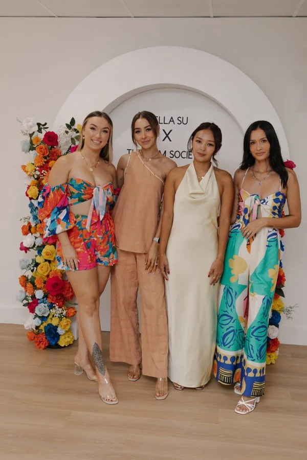 Four women standing in front of a floral display with a white arch and text, dressed in colorful summer outfits, smiling for a photo.