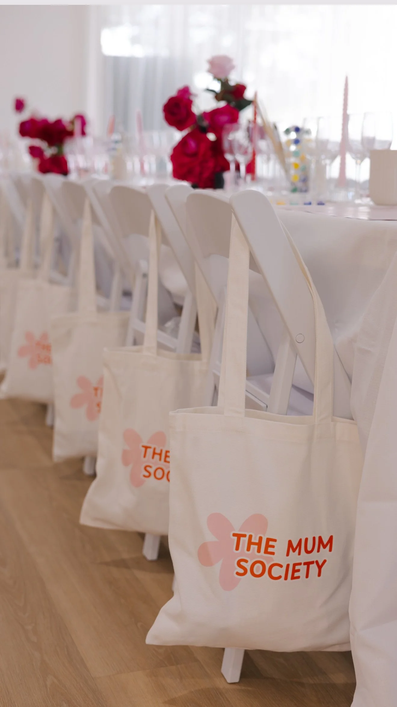 A row of white tote bags with pink flower and orange text 'The Mum Society' hanging on white chairs at a decorated table for an event.