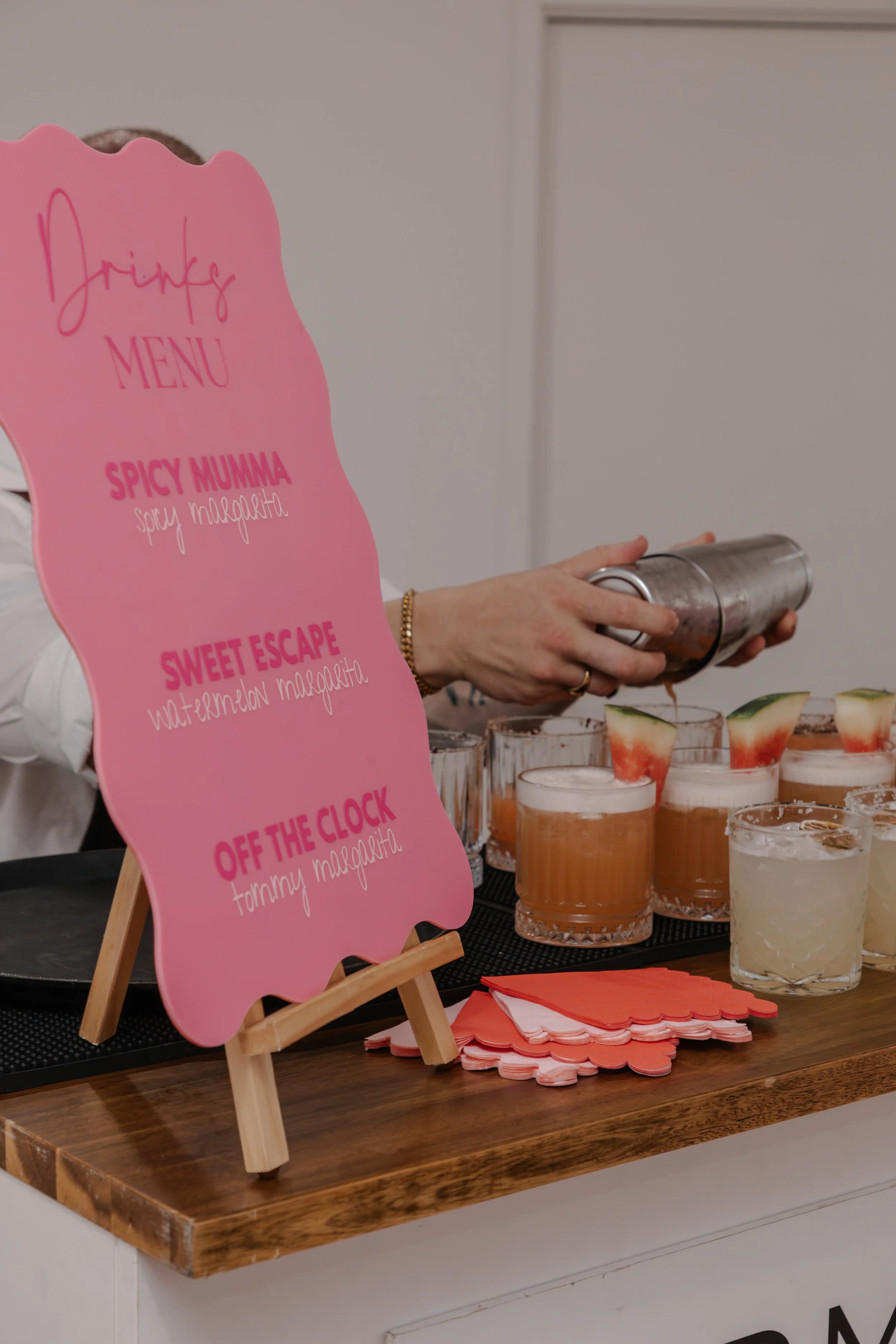 A pink menu board with drink names: Spicy Mumma, Sweet Escape, Off the Clock, with a person pouring a drink into a glass with watermelon garnish behind a table of various drinks.