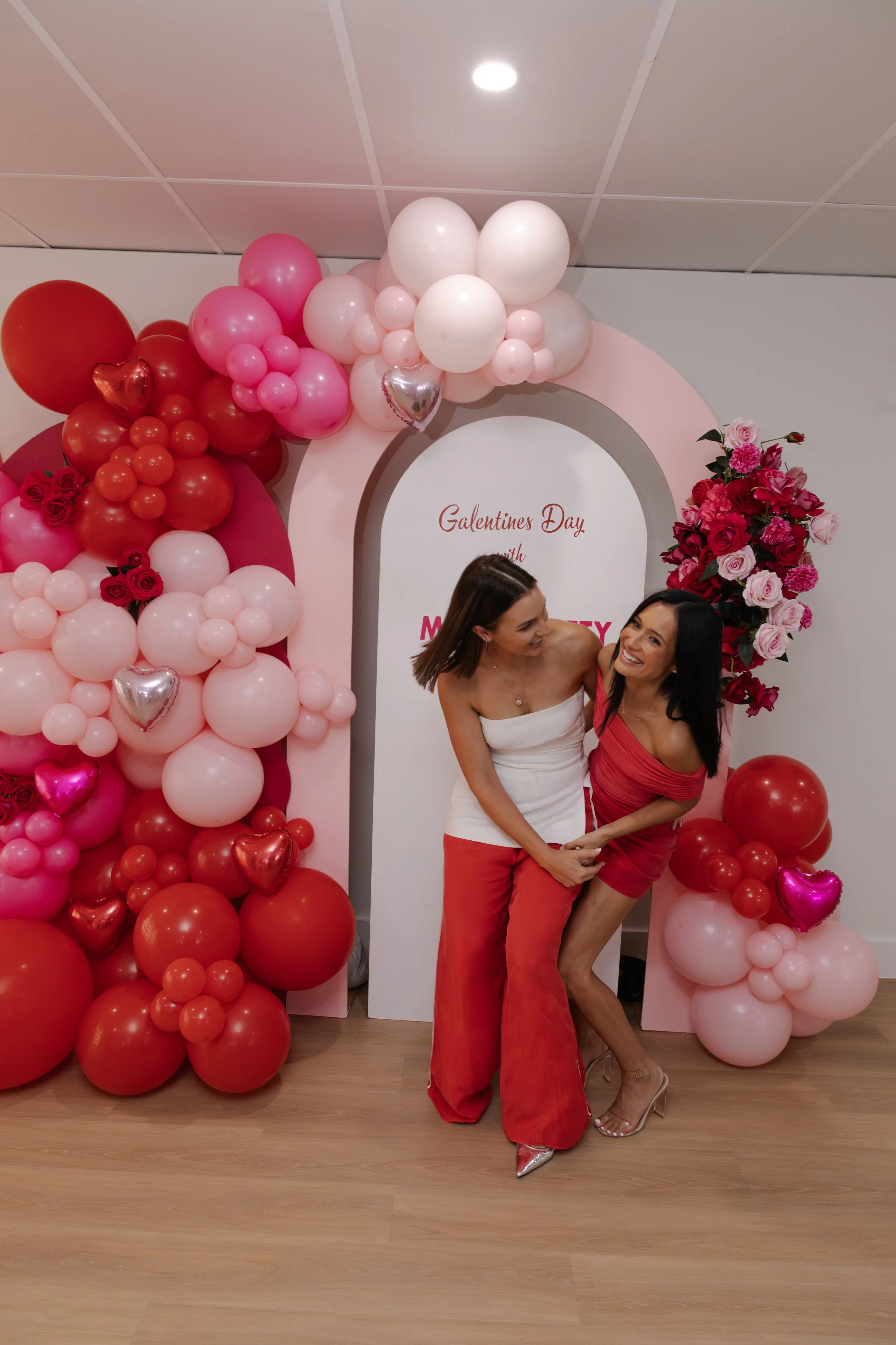 Two women celebrating Valentine's Day indoors, surrounded by pink and red balloons, flowers, and heart-shaped decorations.