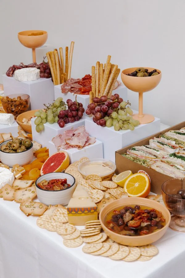 An assortment of cheeses, crackers, fresh grapes, sliced citrus fruits, charcuterie, bread, and spreads on a white covered table, with a variety of snacks and appetizers arranged elegantly.