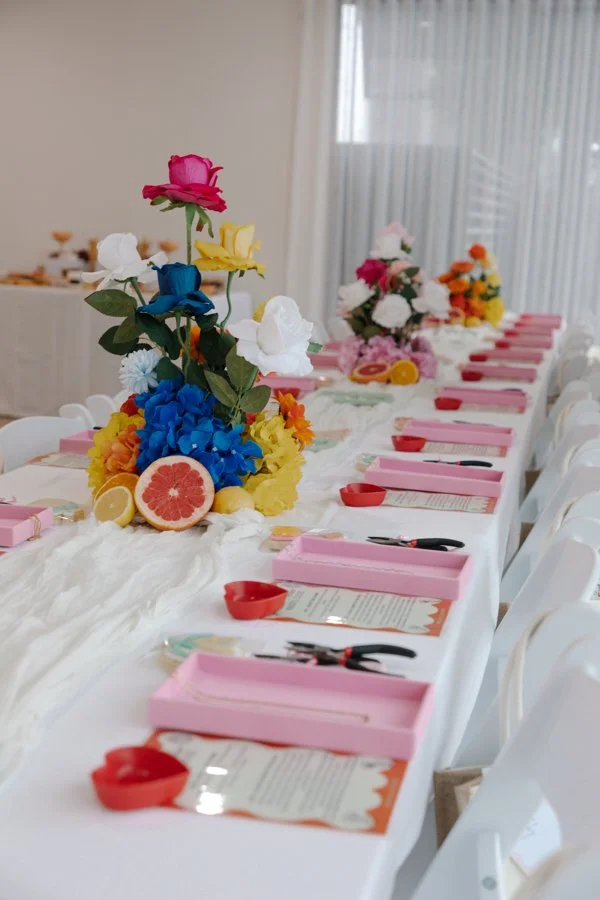Decorated dining table with pink boxes, red bowls, and colorful floral centerpieces including roses, hydrangeas, and citrus slices, set for a celebration or event.