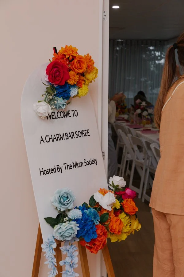 Welcome signboard with colorful artificial flowers for a Charm Bar Soiree hosted by The Mum Society, positioned beside an indoor event space.