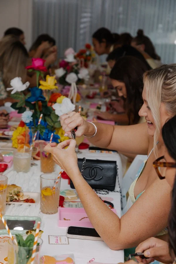 Women sitting at a long table decorated with colorful flowers, enjoying a celebration with food, drinks, and party favors.