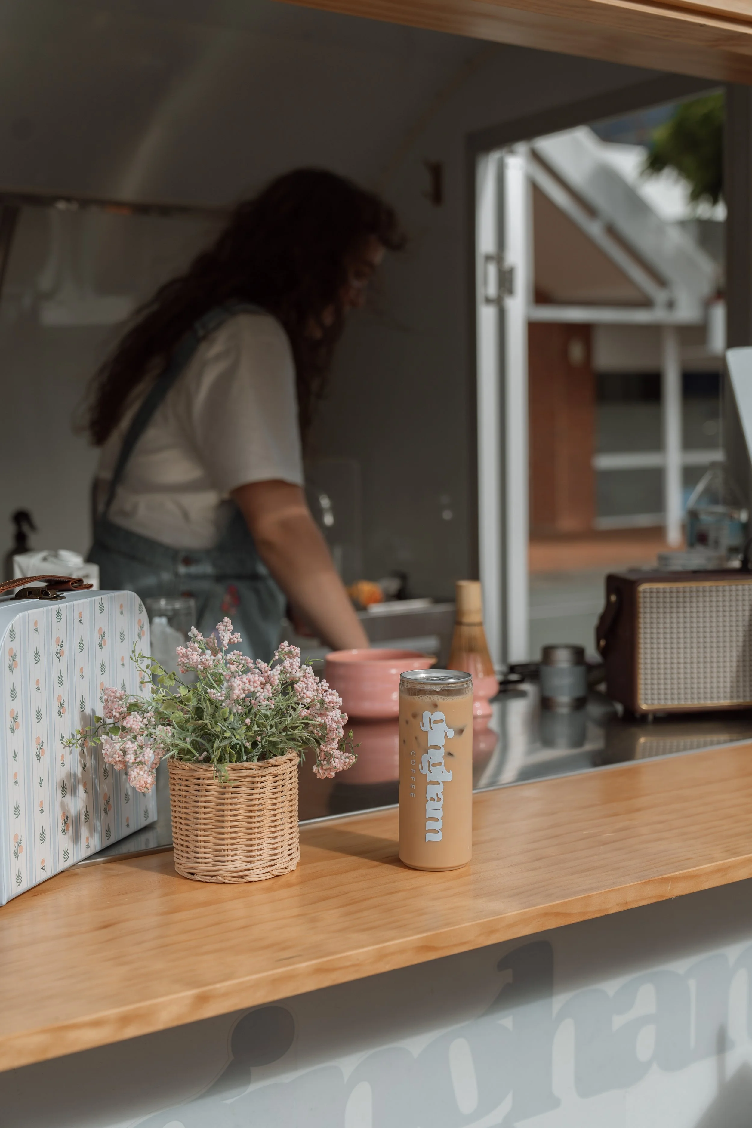 A pink and beige iced coffee in a tall transparent cup with a metal lid, placed on a wooden counter next to a small pink ceramic pot and a woven basket with pink flowers. A woman with curly hair, wearing a white shirt and overalls, is behind the coun