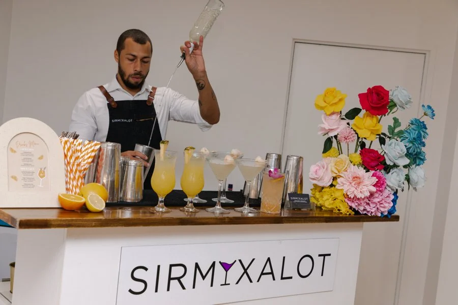 Bartender pouring a cocktail at a bar decorated with colorful artificial flowers and lemons, with a sign that reads 'SIRMXALOT'.
