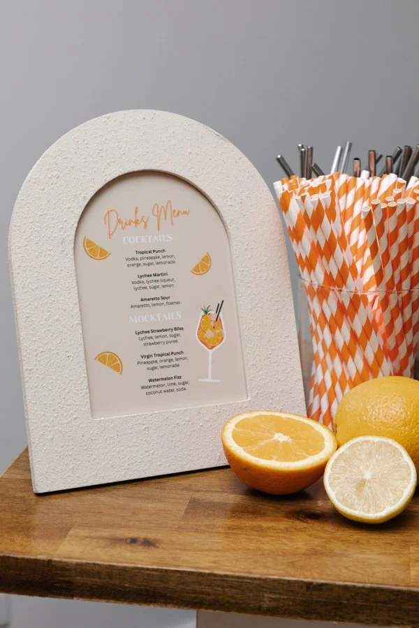 A white arched cocktail menu sign on a wooden table, with a list of drinks, next to a container of orange and white striped straws, and fresh lemons and an orange cut in half.