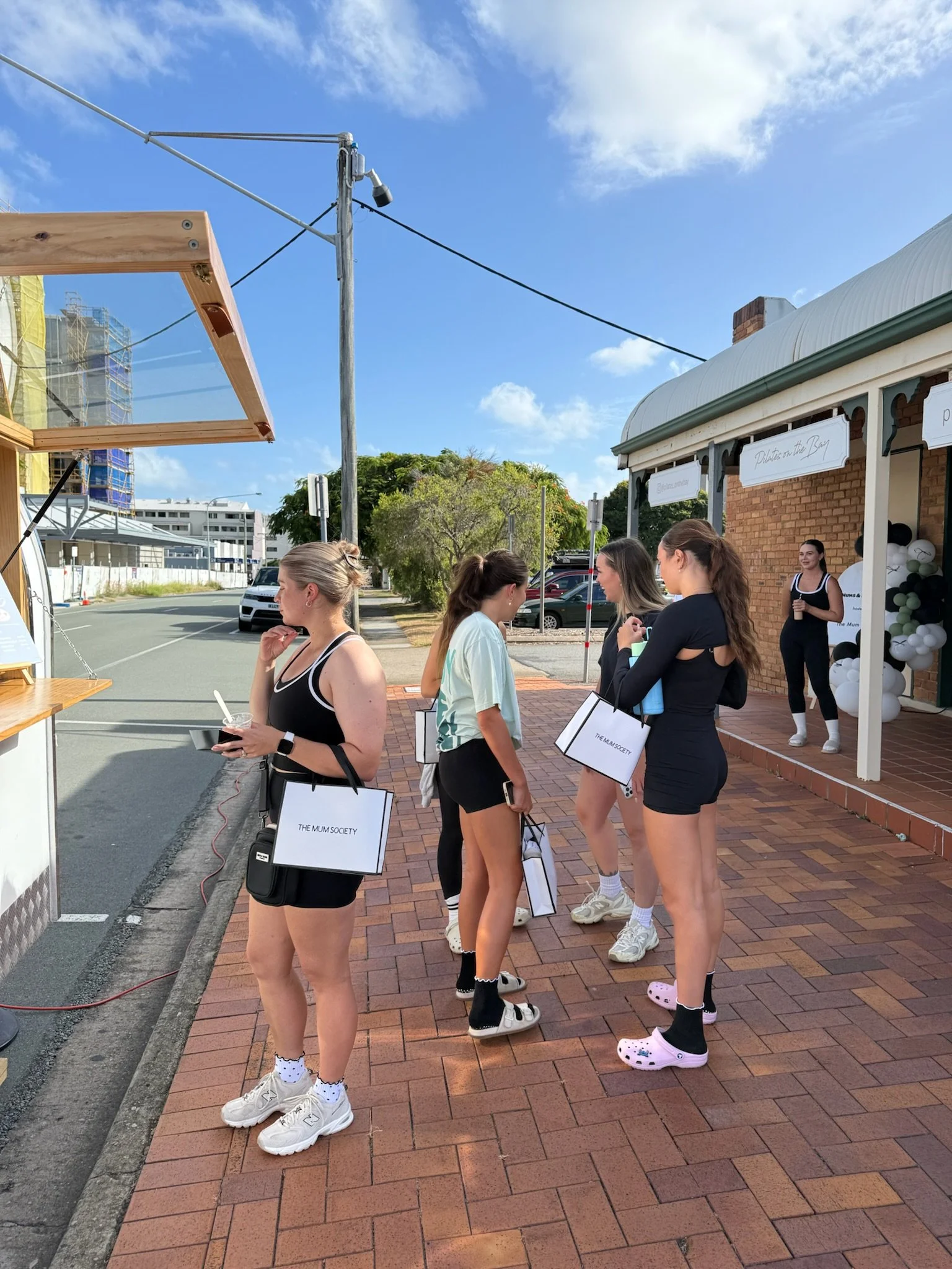 Group of young women casually dressed, standing on a brick sidewalk outside a brick building decorated with balloons, with some holding shopping bags, possibly at an event or gathering.