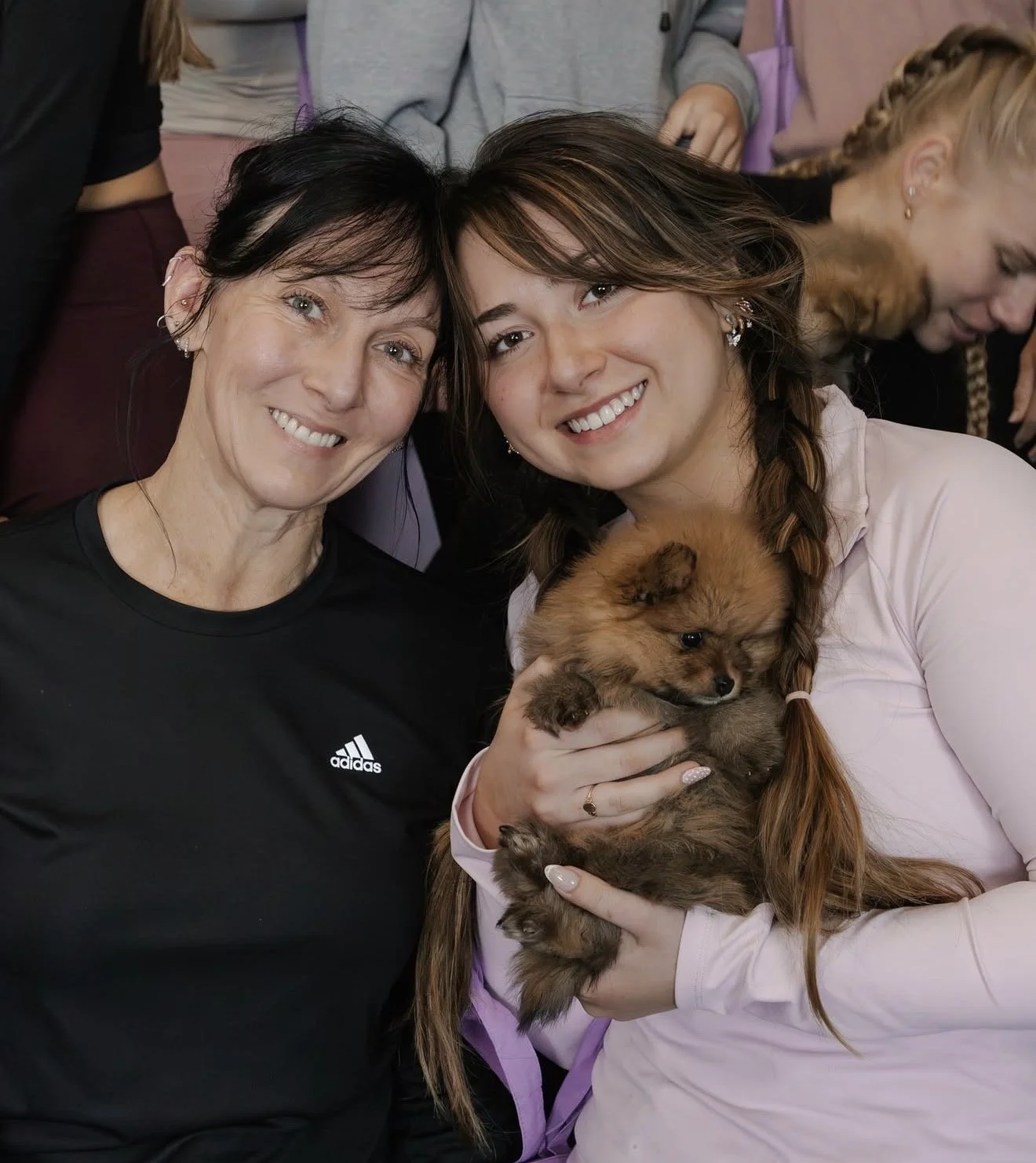 Two women smiling, one holding a small brown puppy, with other people in the background.