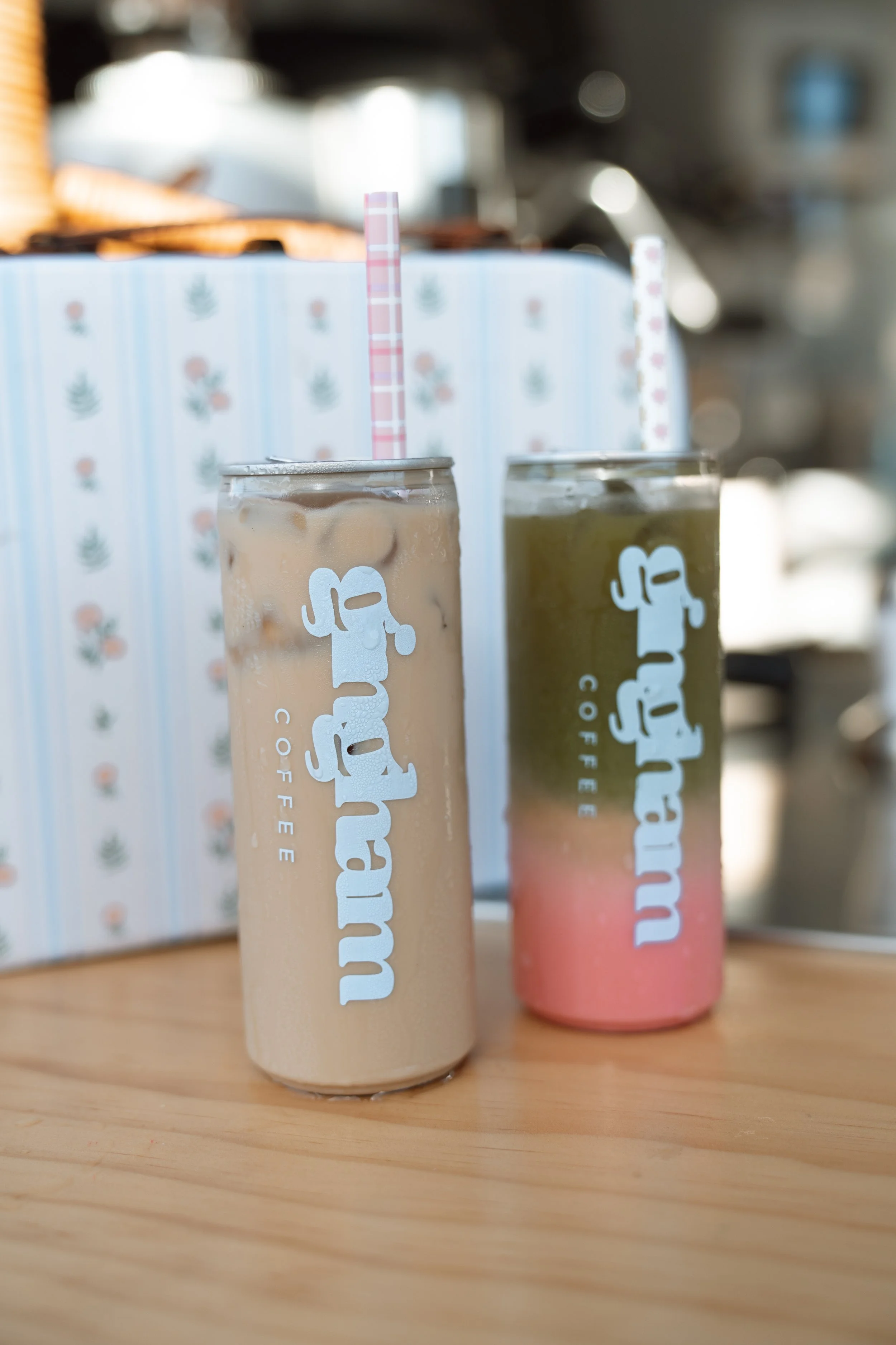 Two colorful iced beverages in tall cans with straws, placed on a wooden surface, with a patterned box in the background.