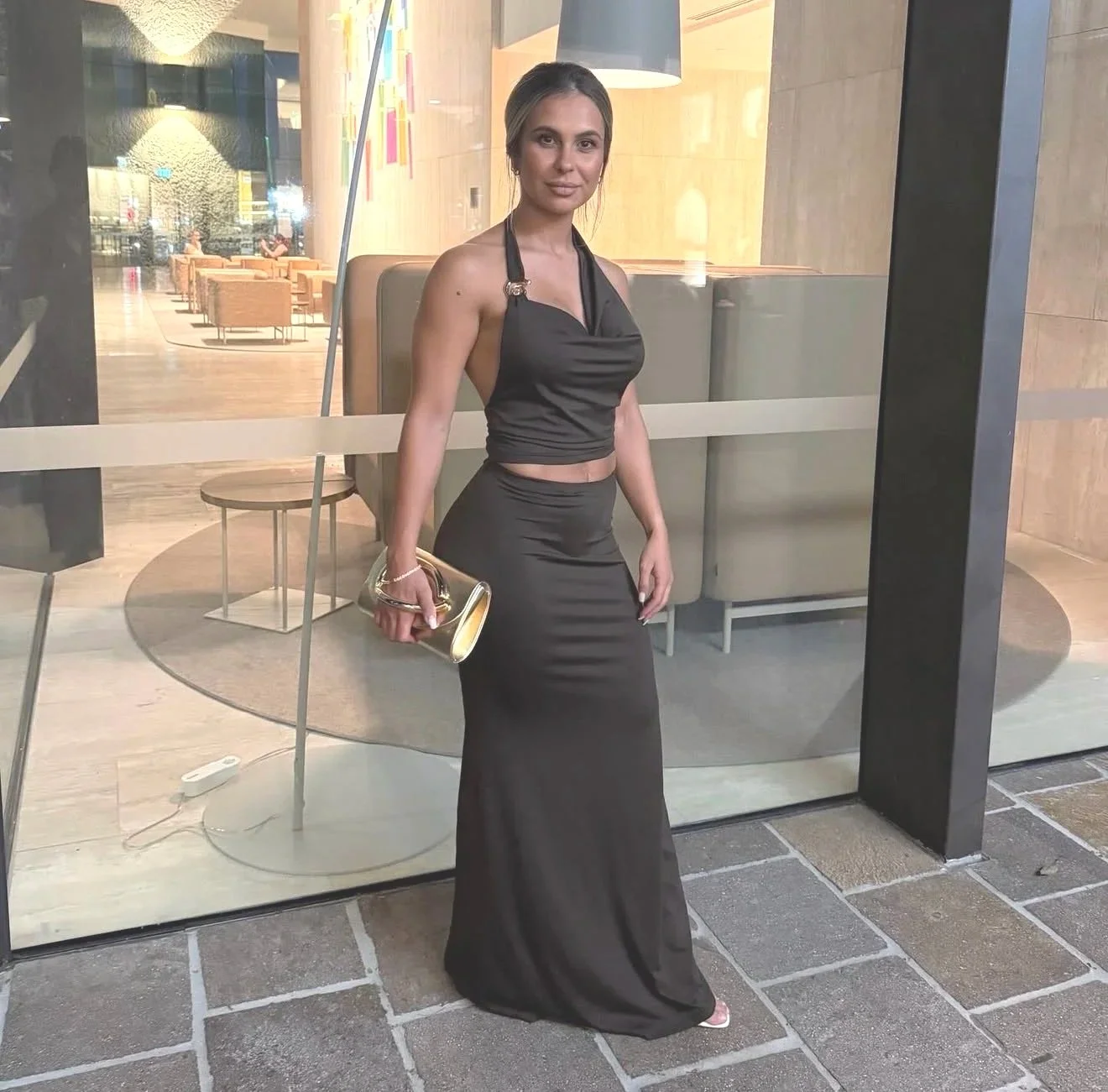 A woman wearing an elegant black evening gown, holding a small gold clutch, standing in front of a glass door at night in a stylish indoor setting.