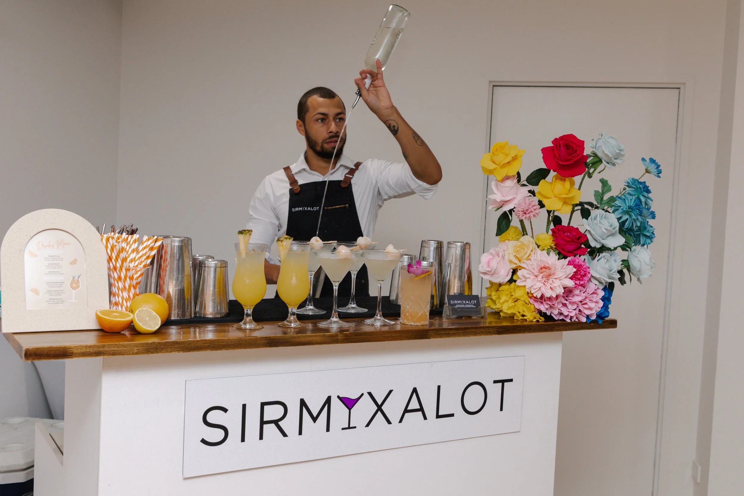 Bartender pouring a drink at a bar decorated with colorful paper flowers and lemon-based cocktails, with a sign reading 'SIRMYXALOT' on the front.