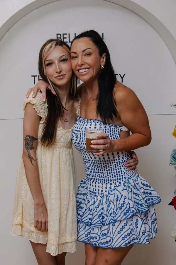 Two women standing close together, smiling, in front of a white wall with text. One woman is holding a drink, wearing a blue and white dress. The other woman is in a light-colored dress with floral details.