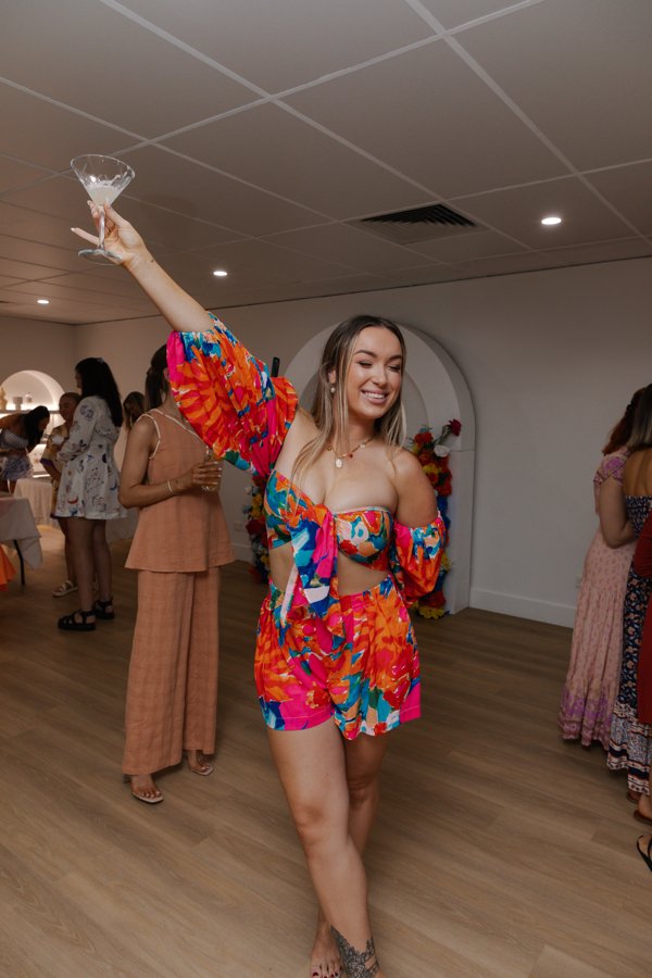 A woman in a colorful floral off-shoulder top and matching shorts holding a martini glass and smiling at a party or celebration.