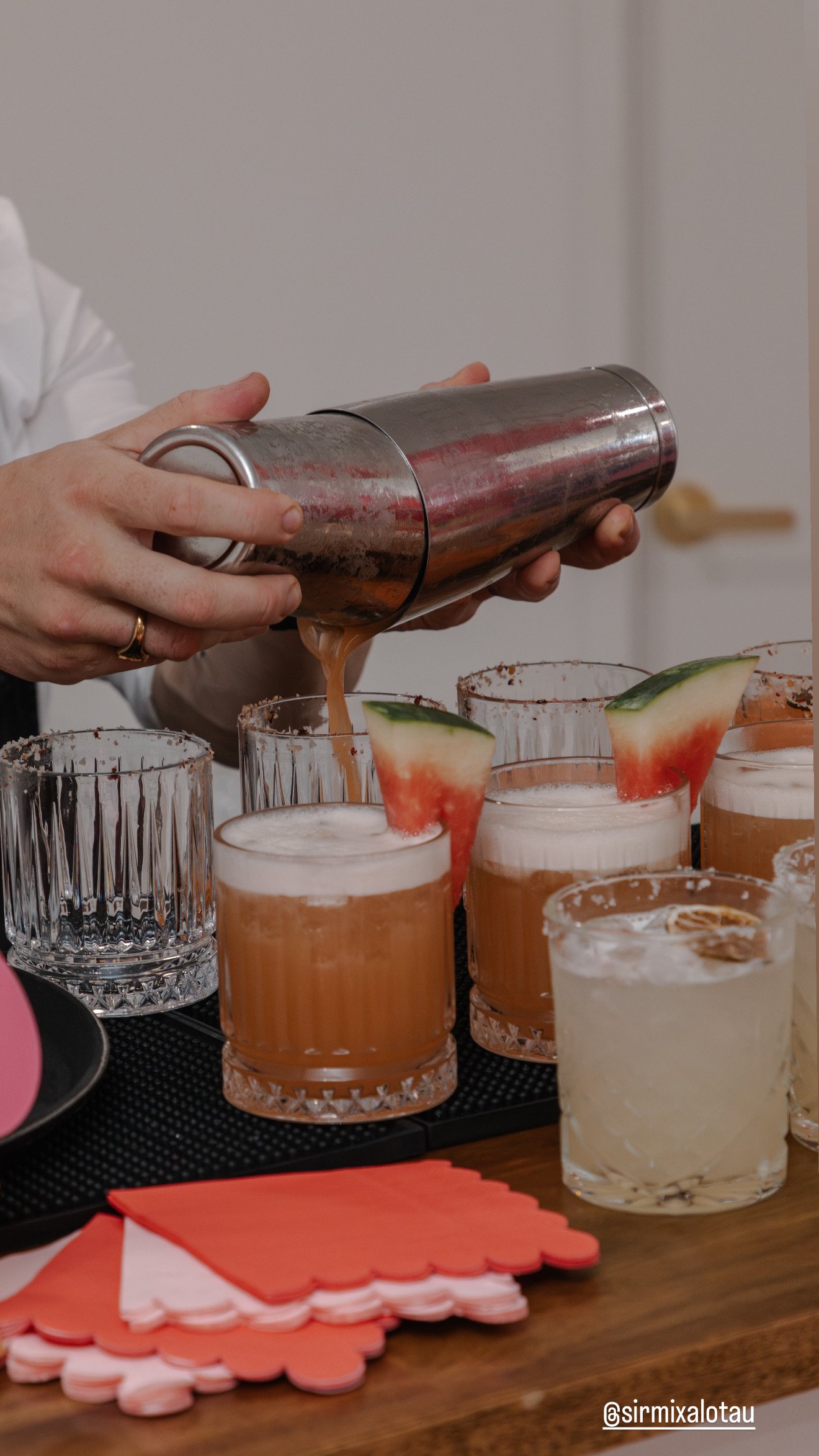 A bartender pours a cocktail into glasses garnished with watermelon slices and cucumber slices.