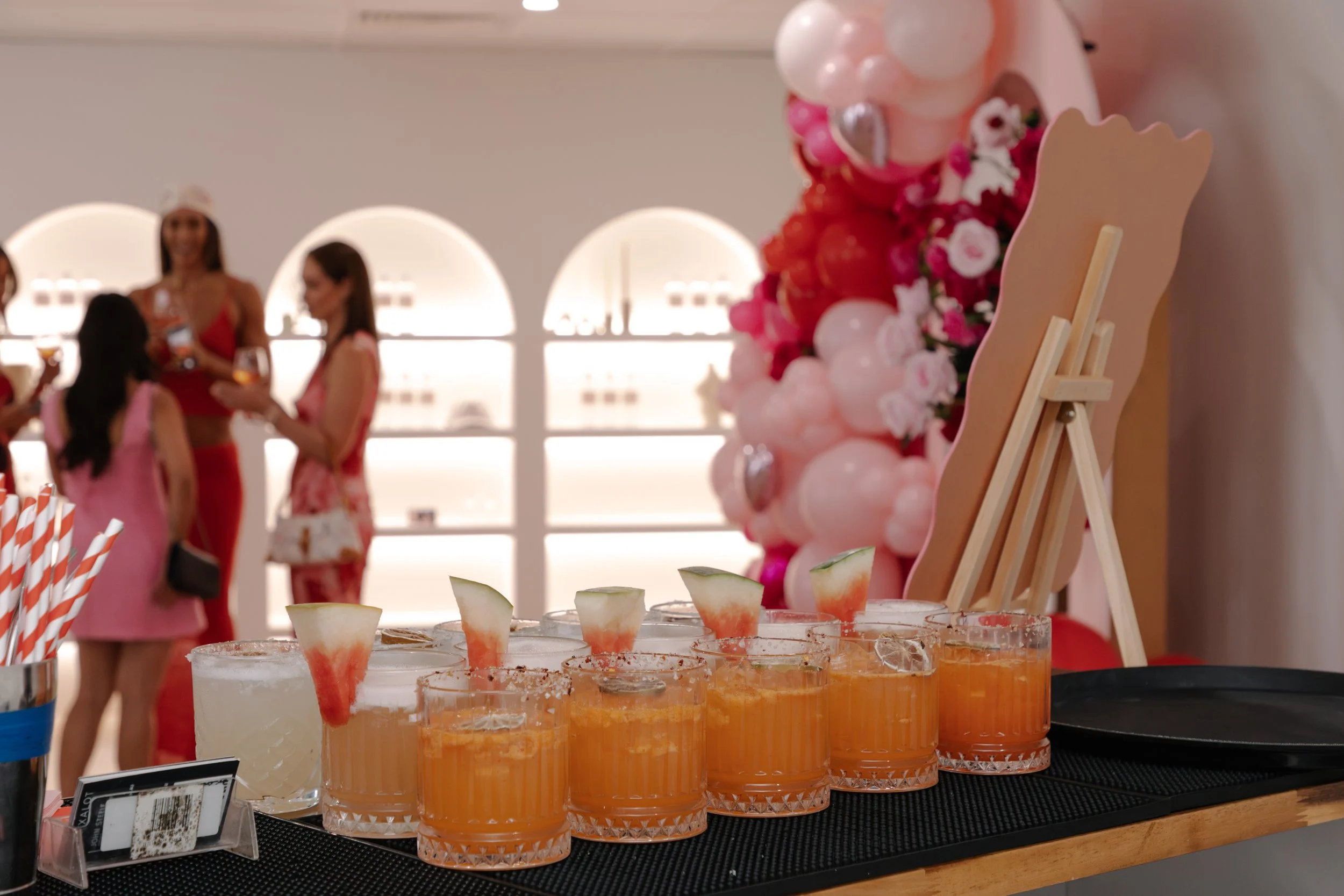 A row of cocktails garnished with watermelon slices on a black bar mat, with pink and red balloon decorations and people socializing in the background.