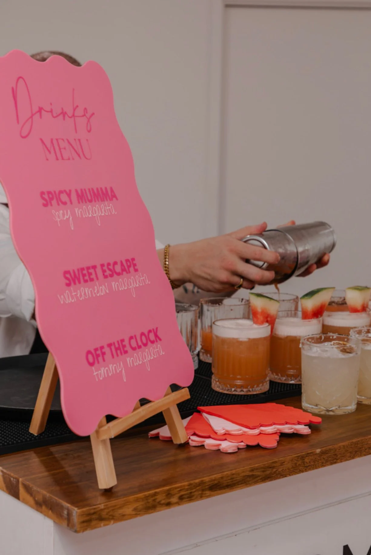 Pink drink menu sign with four beverage options and watermelon garnished drinks on the table.